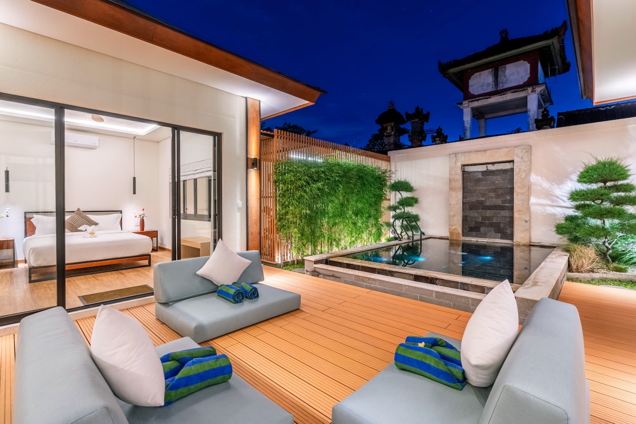 Deck daybeds at dusk, a bedroom lit behind the glass